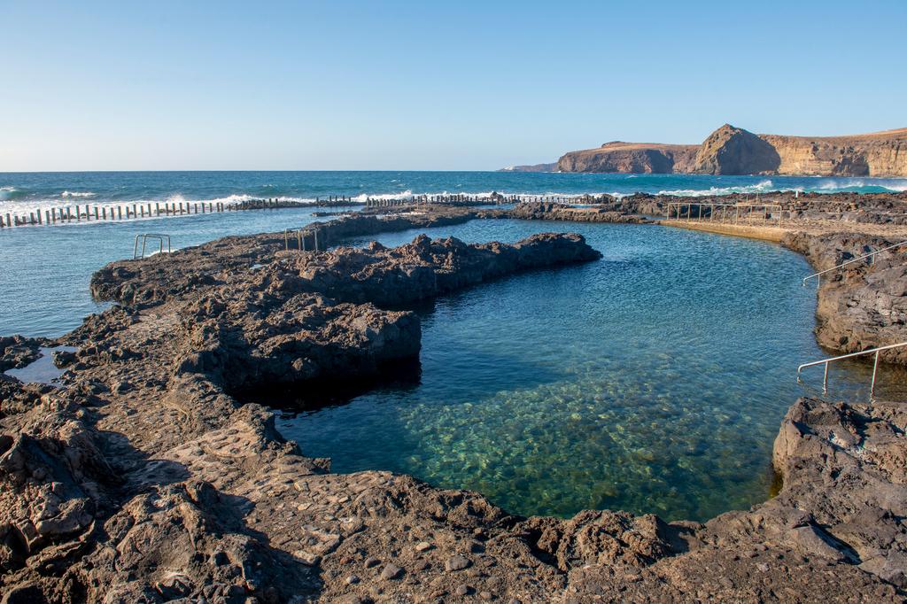Blick von einer Klippe auf den Hafen von Puerto de las Nieves auf Gran Canaria. Im Vordergrund ragen vulkanische Felsen mit grüner Vegetation ins Bild, unten liegt der Hafen mit einem Fährschiff und weißen Häusern. Dahinter erstreckt sich die Küste bis zum Horizont, umgeben vom blauen Atlantik im warmen Abendlicht.